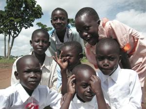 Seven young Black African children cluster up and pose for the camera on a dirt road near tall, leafy trees. The boys wear crisp button-down shirts, and the girls where pastel dresses. All have very short hair.